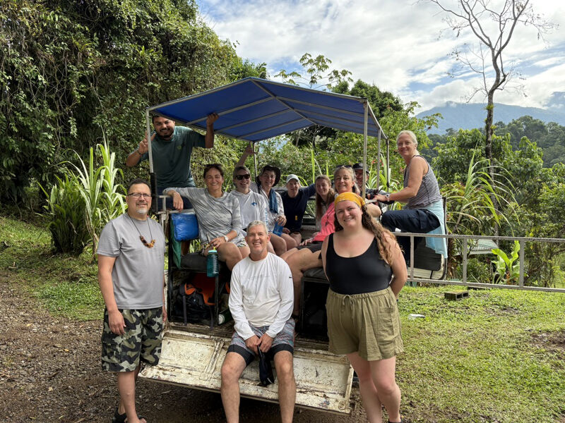A group of people is posing for a photo in the back of a truck. The truck has a blue canopy. They are surrounded by lush greenery, suggesting they are in a natural environment. The people appear to be enjoying themselves, smiling at the camera. The setting seems to be a sunny day.